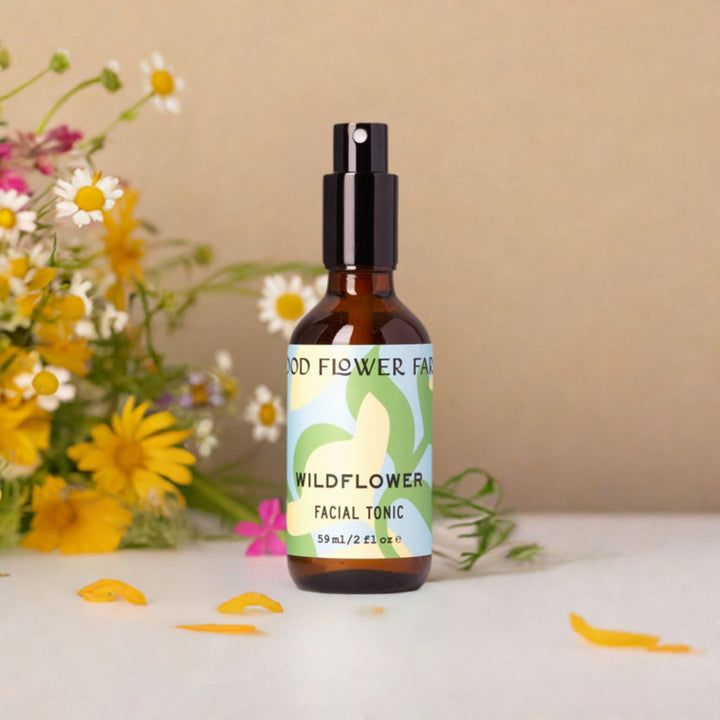 Wildflower Facial Tonic in amber bottle on white table, surrounded by yellow petals and wildflower stems.