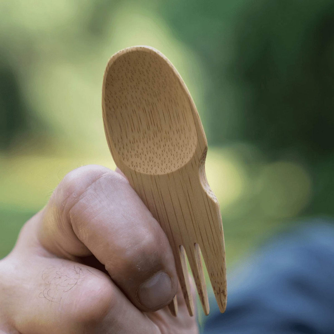 Bamboo spork in hand with blurred clothing and greenery in background.