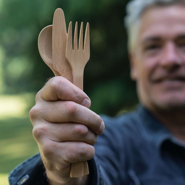 Bamboo utensil set, including spoon, knife, and fork shown in hand with man and greenery in background.  