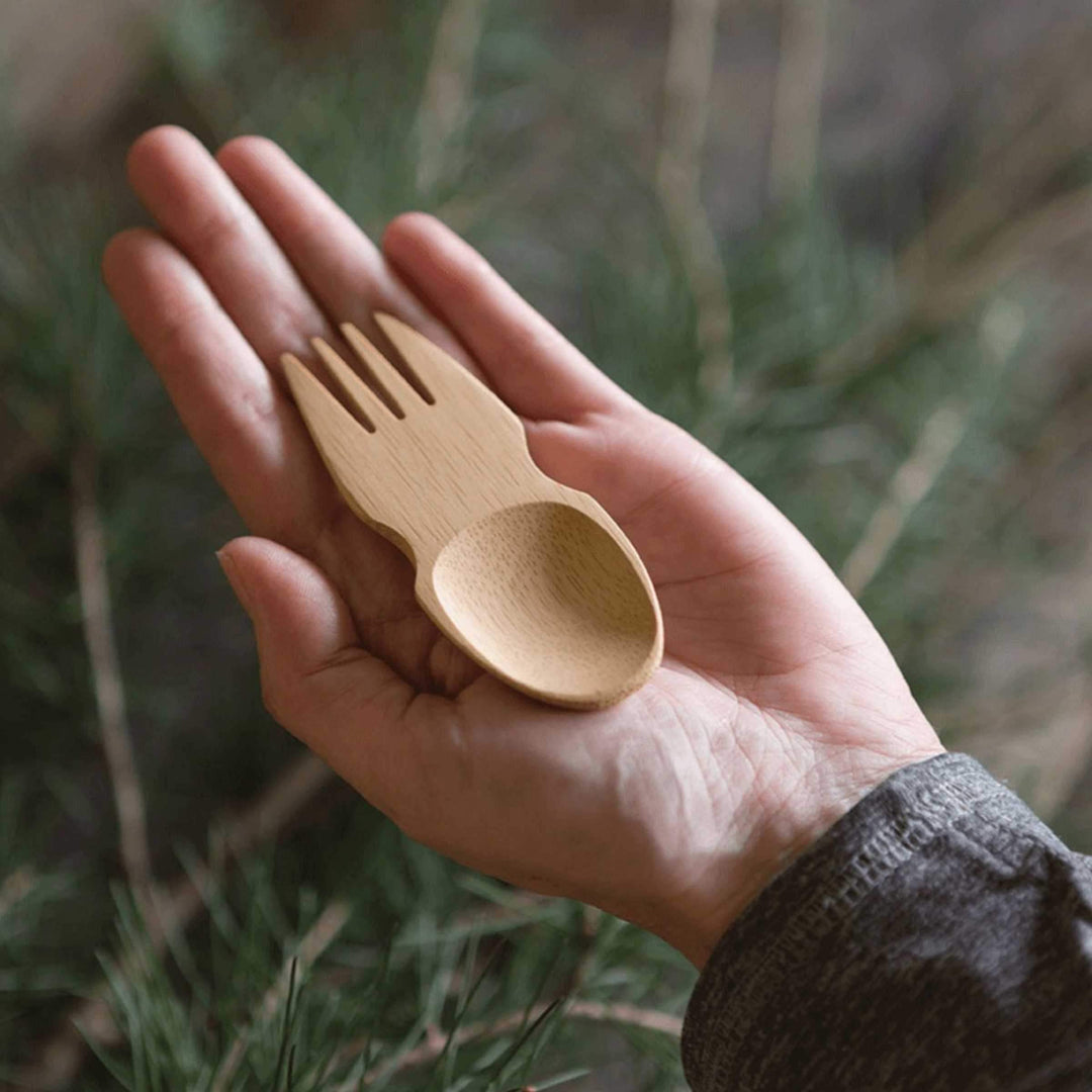 Single bamboo spork resting in person's palm with  evergreen tree in background.