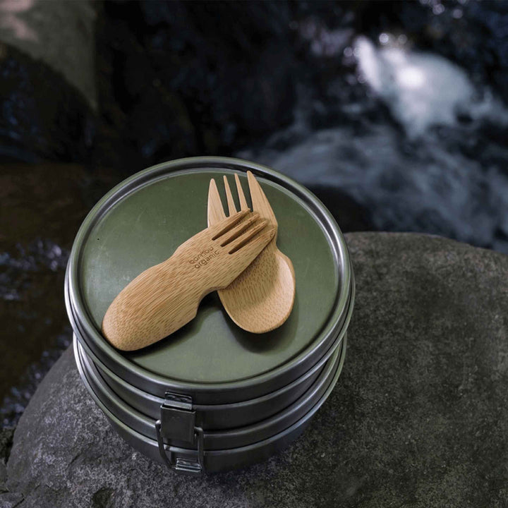 Two bamboo sporks atop stainless steel food container set on rock with waterfall in background.