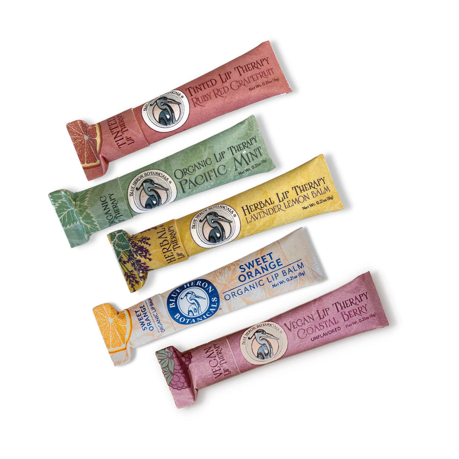 Blue Heron Botanical Lip Therapy Balms in 5 formulas from top: Ruby Red Grapefruit (tinted), Pacific Mint, Lavender Lemon, Sweet Orange, and Coastal Berry (unflavored), packaged in compostable paper tubes.