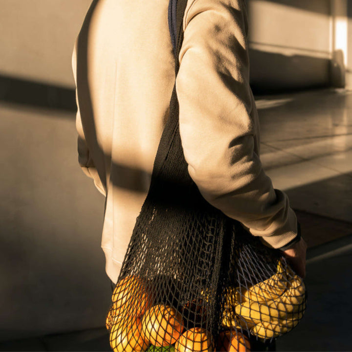 Ecobags String Bags in Black color, shown with oranges and bananas, over shoulder of person with camel colored coat. 