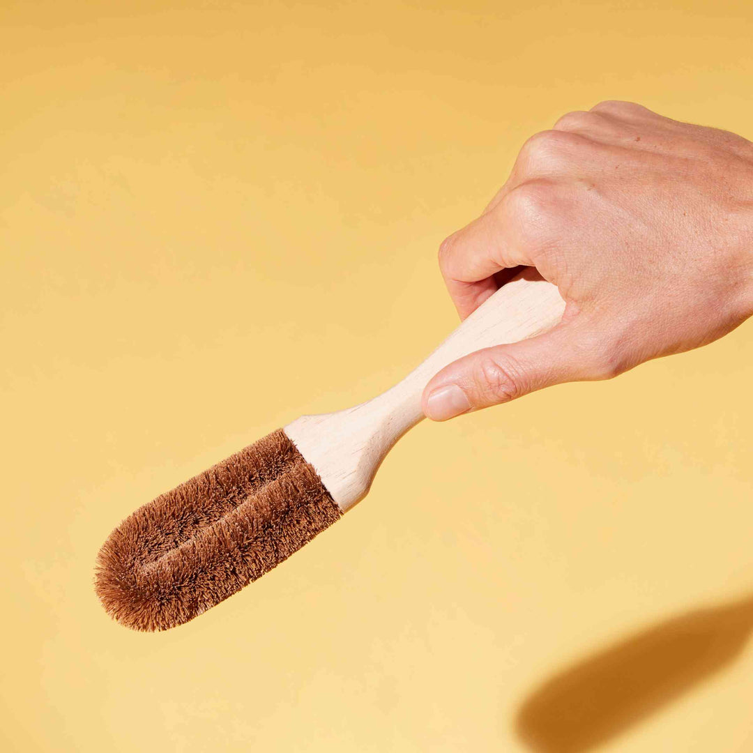 Ecococonut Dish Brush held in person's hand with yellow background.