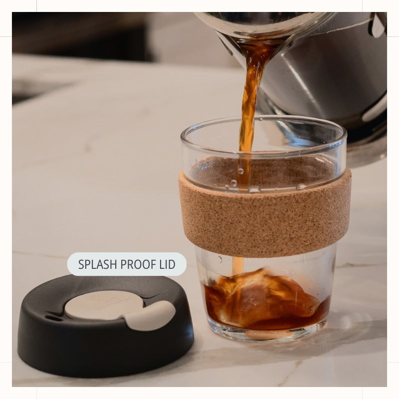 Glass Coffee Cup with Lid and Cork Band in Press/Charcoal color with coffee pouring from French press. Note indicates splash-proof lid.