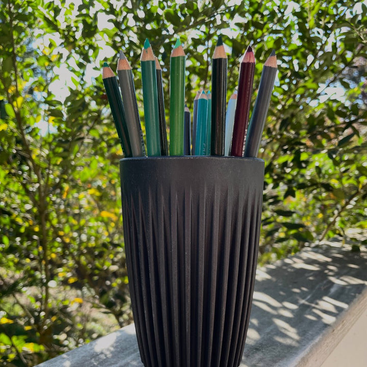 HuskeeCup and lid in charcoal color, shown repurposed as a pencil holder on white railing with bushes in background.