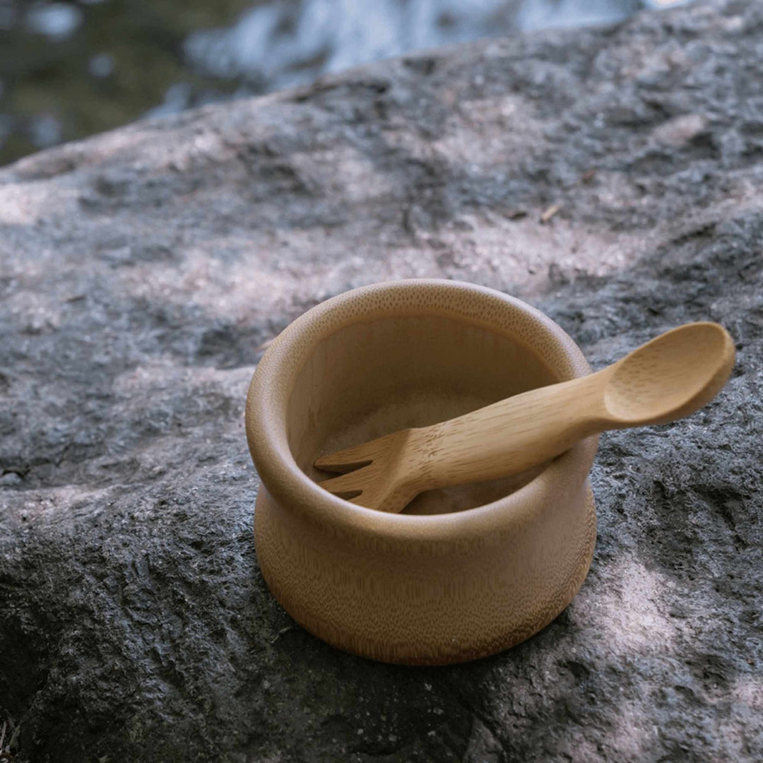 Kid's bamboo spork, shown in small bowl on rock with stream in background.