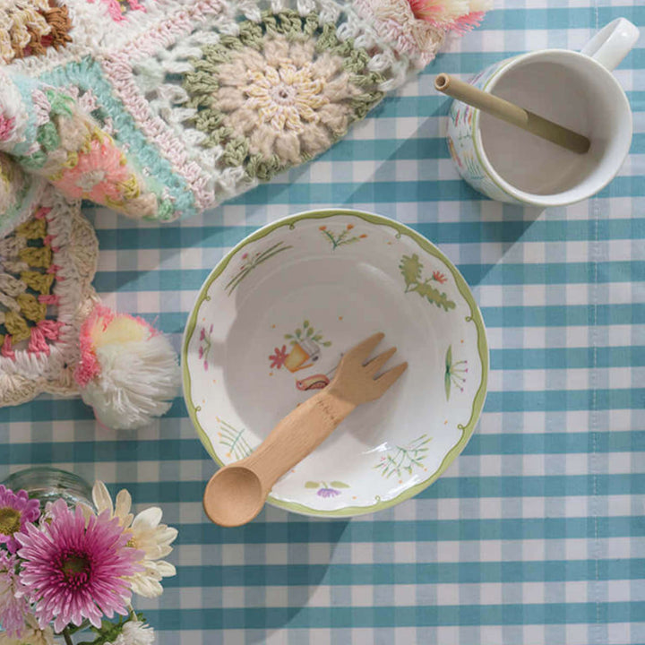 Kid's bamboo spork, shown in decorative bowl with mug, crocheted blanket and flowers in small vase on blue and white checkered tablecloth.
