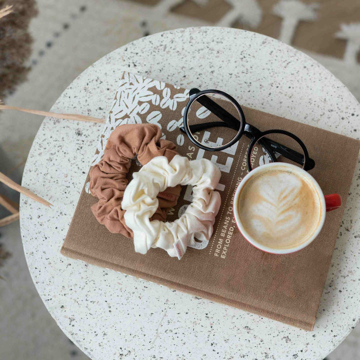 Cappuccino hair scrunchies with a book, eyeglasses, and a coffee drink set on small table.