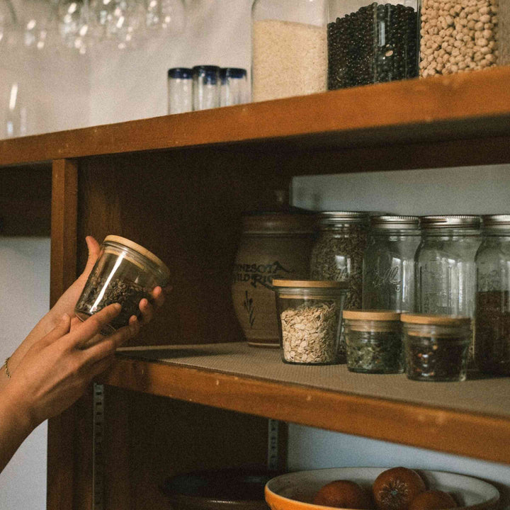 Bamboo Mason jar lids shown on glass jars with food storage on pantry shelf with hands holding one jar. Other glass canisters in background.