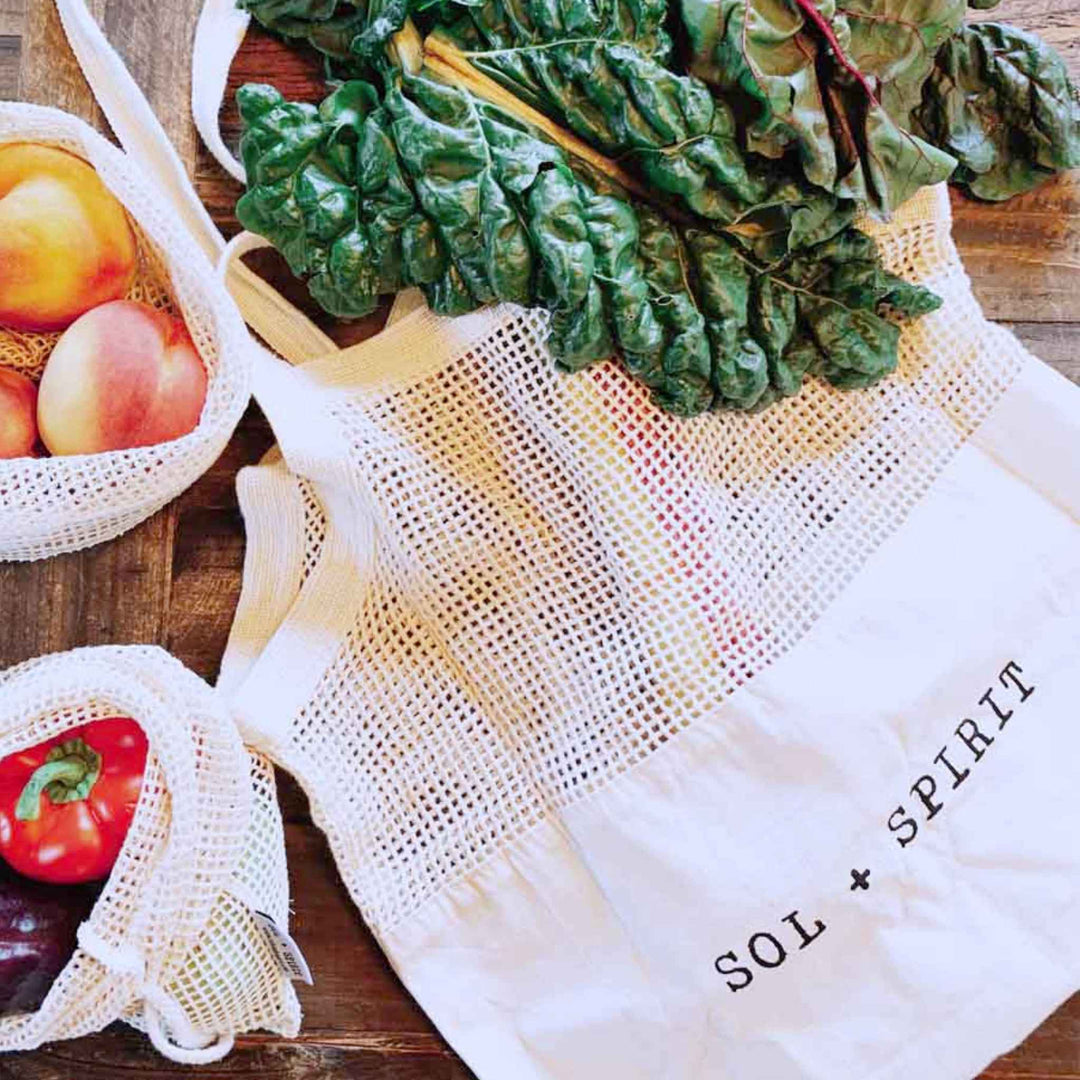 SOL + SPIRIT Organic Cotton Reusable Mesh And Canvas Sling Shopping Bag Laid Flat on Wood Table with Fruit and Greens Surrounding the Bag.