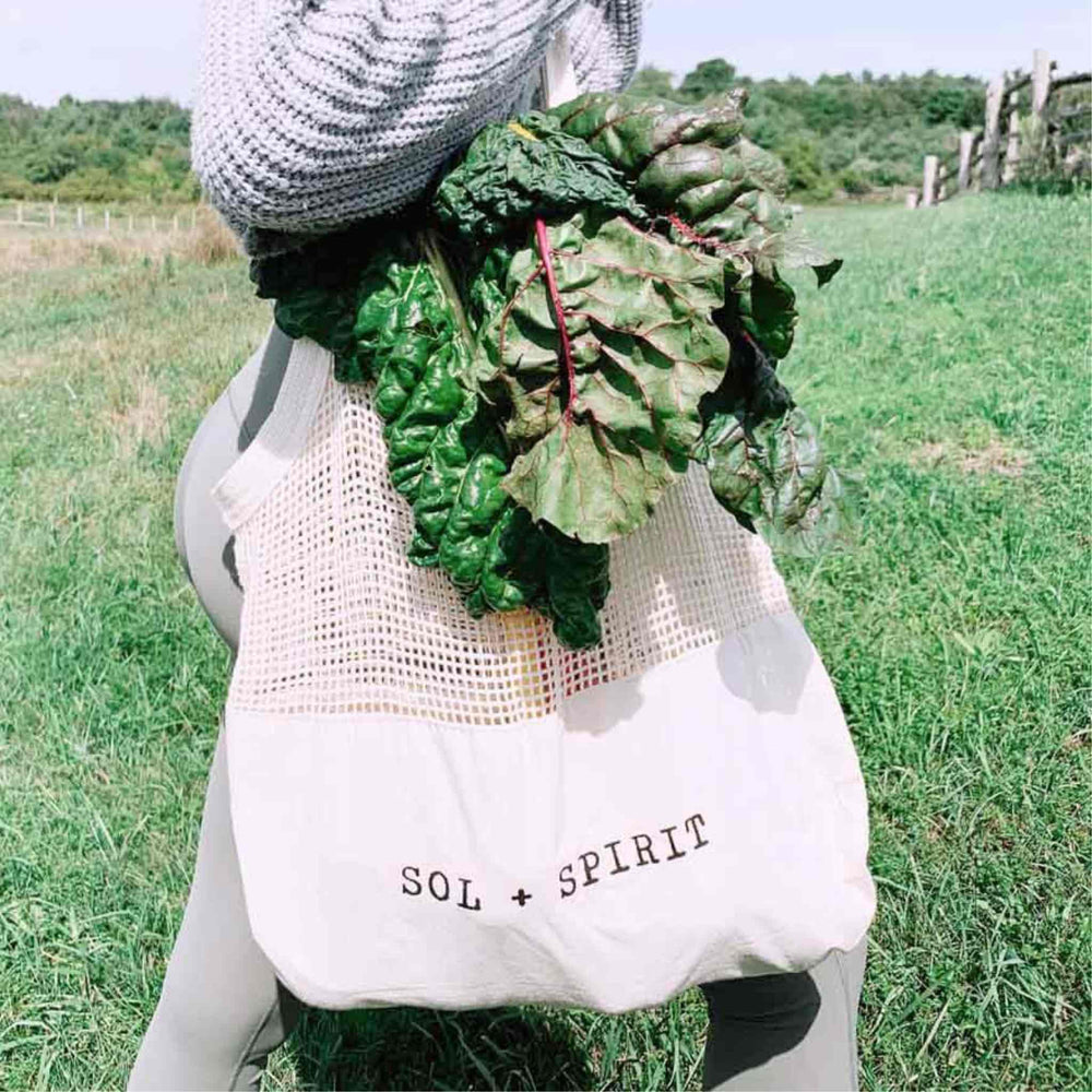 SOL + SPIRIT Organic Cotton Reusable Mesh And Canvas Sling Shopping Bag on Woman's Arm with Greens in Bag. Green Grass Field in Background.
