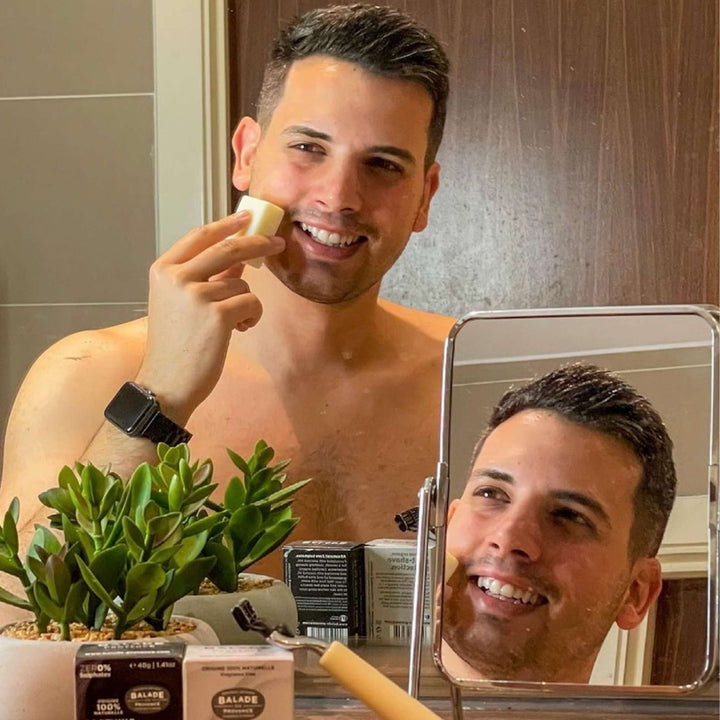 Man using handmade, cold process, vegan shave bar for men in large and small bathroom mirrors, with razor, soap bars in packaging, and plant in foreground.
