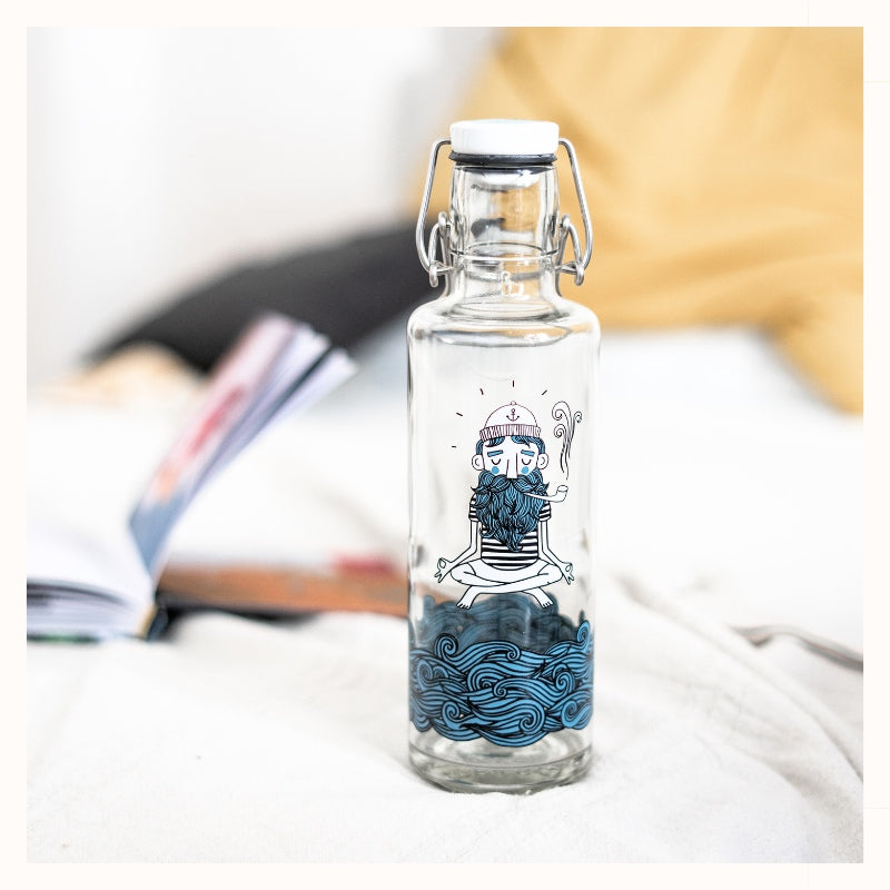 Plastic-free, 20 ounce glass water bottle in Soulsailor design, on bed with book and pillows in background.