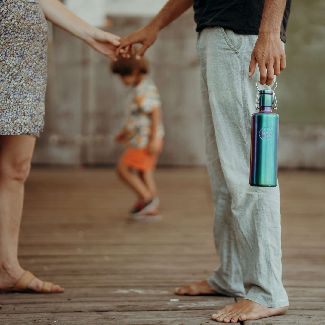 Soulbottles stainless steel light bottle in Utopia color (multi), 25 ounce capacity. held in one hand, while holding other hand with pregnant partner. Child walking in background.