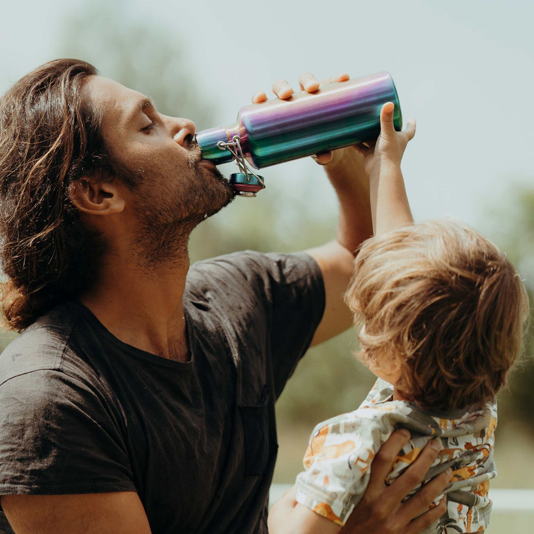 Soulbottles stainless steel light bottle in Utopia color (multi), 25 ounce capacity, in man's hand while drinking. Child is helping tip bottle.
