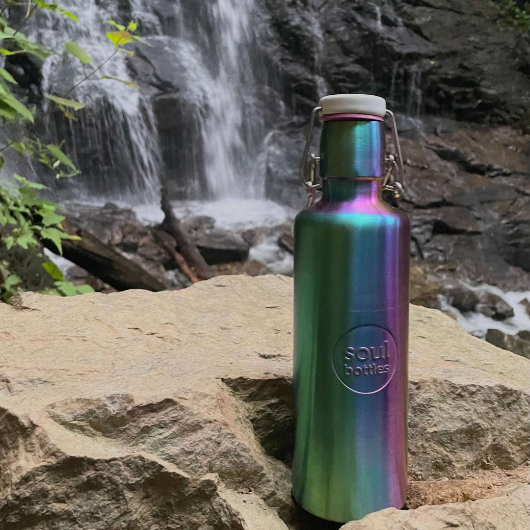 Soulbottles stainless steel light bottle in Utopia color (multi), 25 ounce capacity, resting on rocks with waterfall in background.