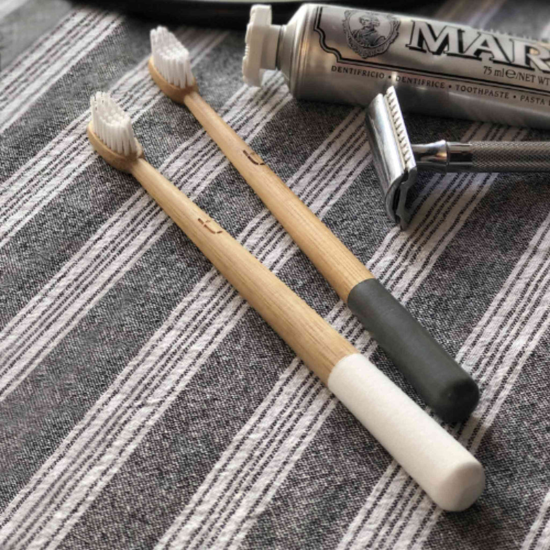 Two Bamboo Toothbrushes for Adults, shown in Cloud White and Storm Gray handle colors on white and gray striped cloth with toothpaste tube and safety razor.