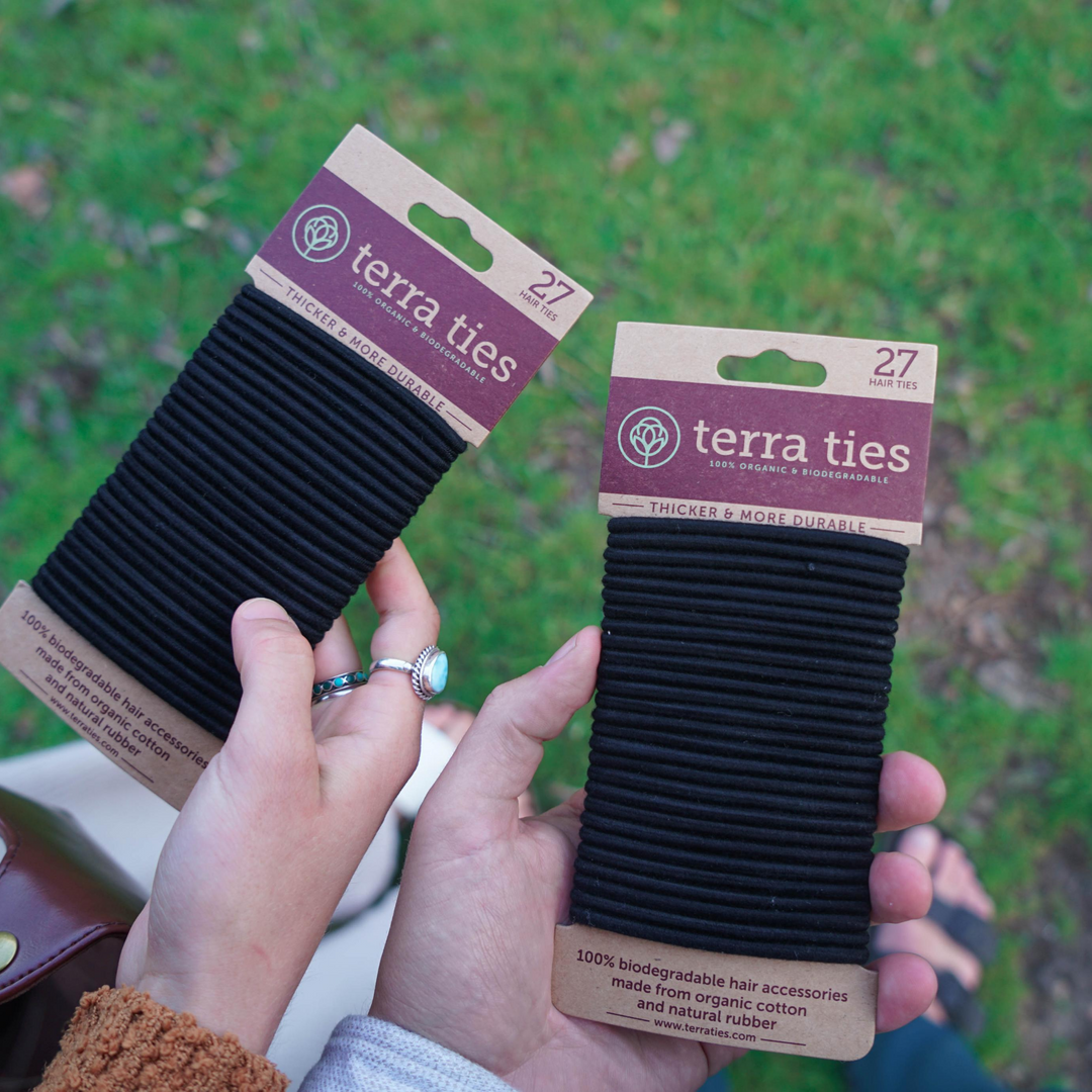 Terra Ties Plastic Free Hair Ties In Black Color. Woman And Man Holding One Pack Each Of 27 Hair Ties On Paper Packaging, With Green Grass In Background.