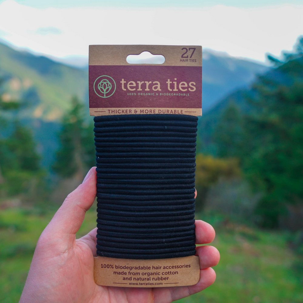 Terra Ties Plastic Free Hair Ties In Black Color. Woman Holding  One Pack Of 27 Hair Ties On Paper Packaging, With Mountains In Background.