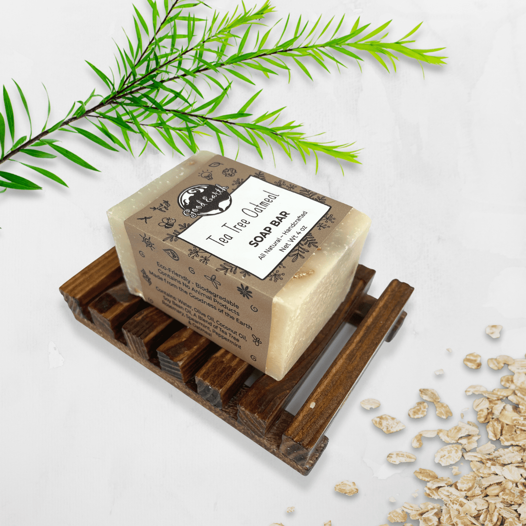 Good Earth Tea Tree Oatmeal Soap Bar In Simple Sleeve Packaging Atop Wood Soap Dish (Sold Separately). Shown With Tea Tree Branch And Loose Oats.
