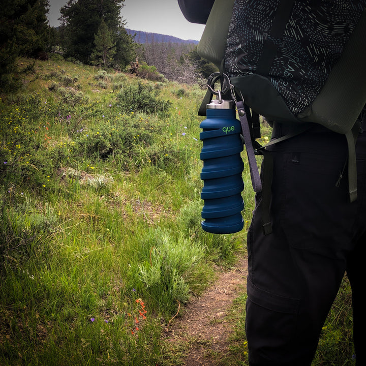 Que Bottle Featuring Bottle Cap With Loop And Multifunctional Keychain. Shown Here Clipped To Backpack, With Hiking Trail In Background.