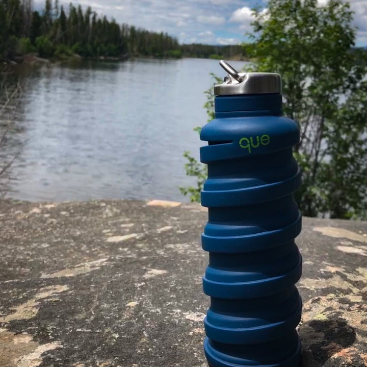 Que Bottle Set On Rock With Lake And Trees In Background